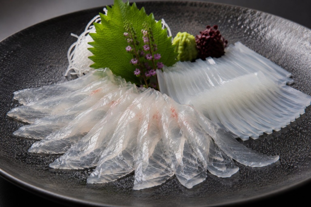 刺身包丁で薄く切った白身魚とイカの刺身を、黒い皿に上品に盛り付けた写真。透き通るような切り身と奥の大葉やつまが見える。
